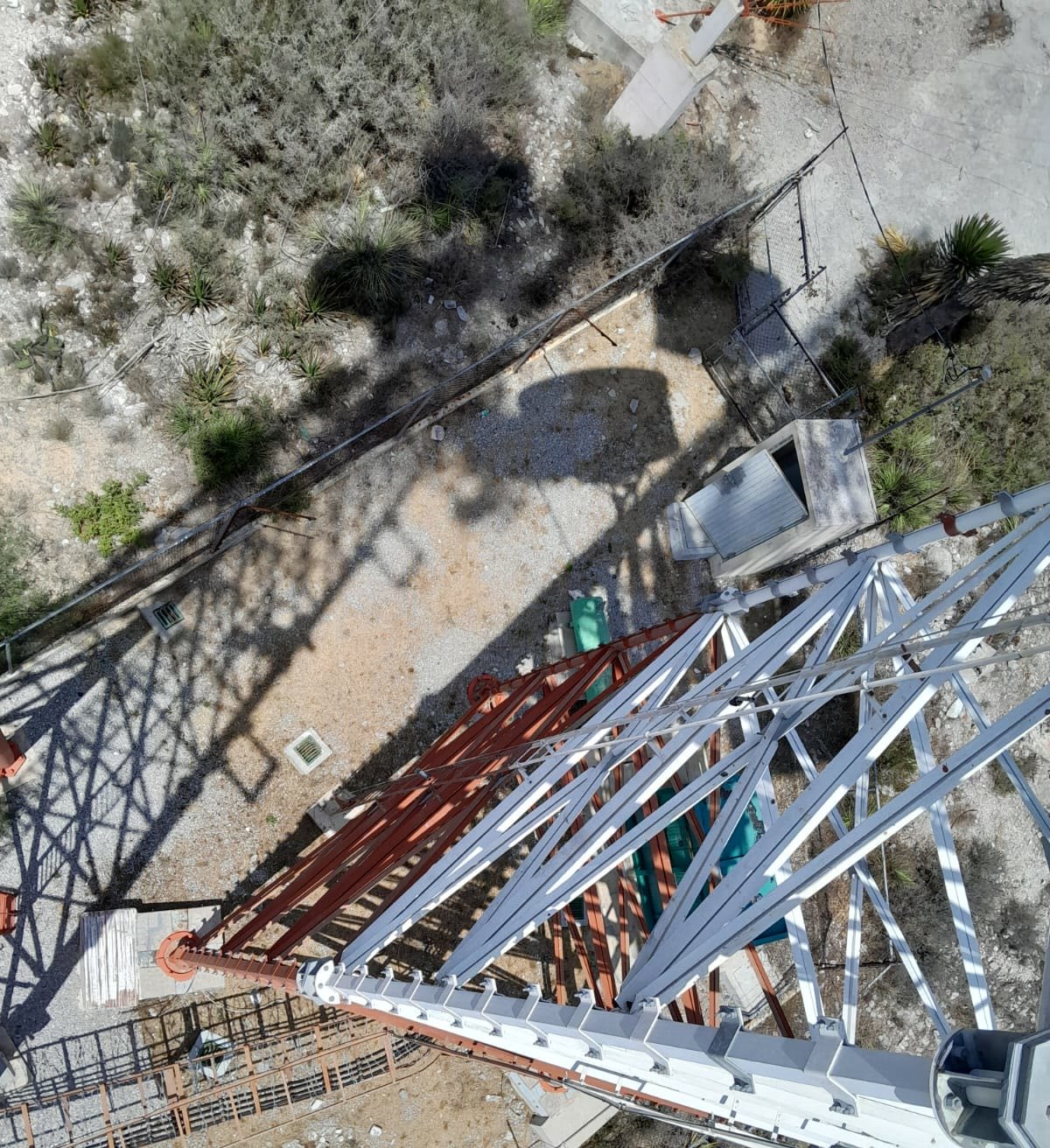 Vista en picada desde lo alto de una torre autosoportada de celosía: los montantes de acero blanco y rojo se abren en perspectiva sobre el terreno semiárido, con la sombra de la propia torre proyectada en el suelo